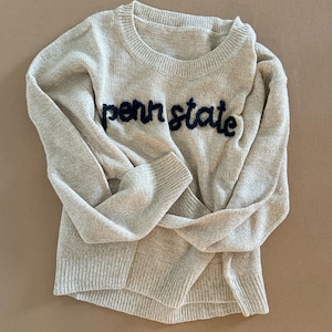 May include: A beige knitted jumper with a black embroidered "penn state" logo.