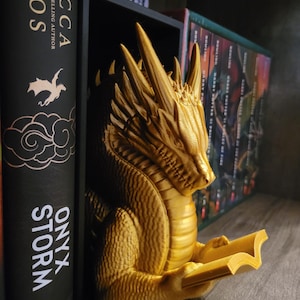 Puede incluir: Un sujetalibros de dragón dorado con un libro en sus garras. El dragón está detallado con escamas y tiene una expresión feroz. El sujetalibros está en un estante con otros libros.