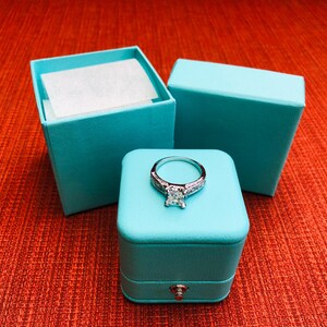 Op de afbeelding: Een turquoise ringdoos met een scharnierend deksel en een zilveren ring met een grote vierkante diamant in het midden.