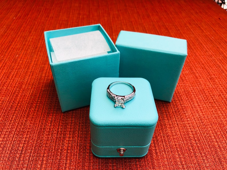 Op de afbeelding: Een turquoise ringdoos met een scharnierend deksel en een zilveren ring met een grote vierkante diamant in het midden.