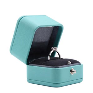 Op de afbeelding: Een turquoise blauwe ringendoos met een zwart fluwelen interieur. De doos is open en een zilveren ring met een enkele diamant zit erin.