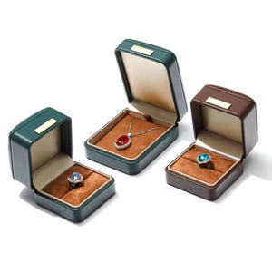 May include: Three green and brown velvet jewelry boxes with lids open. The boxes are lined with brown velvet and contain a red gemstone pendant, a blue gemstone ring, and an empty space.