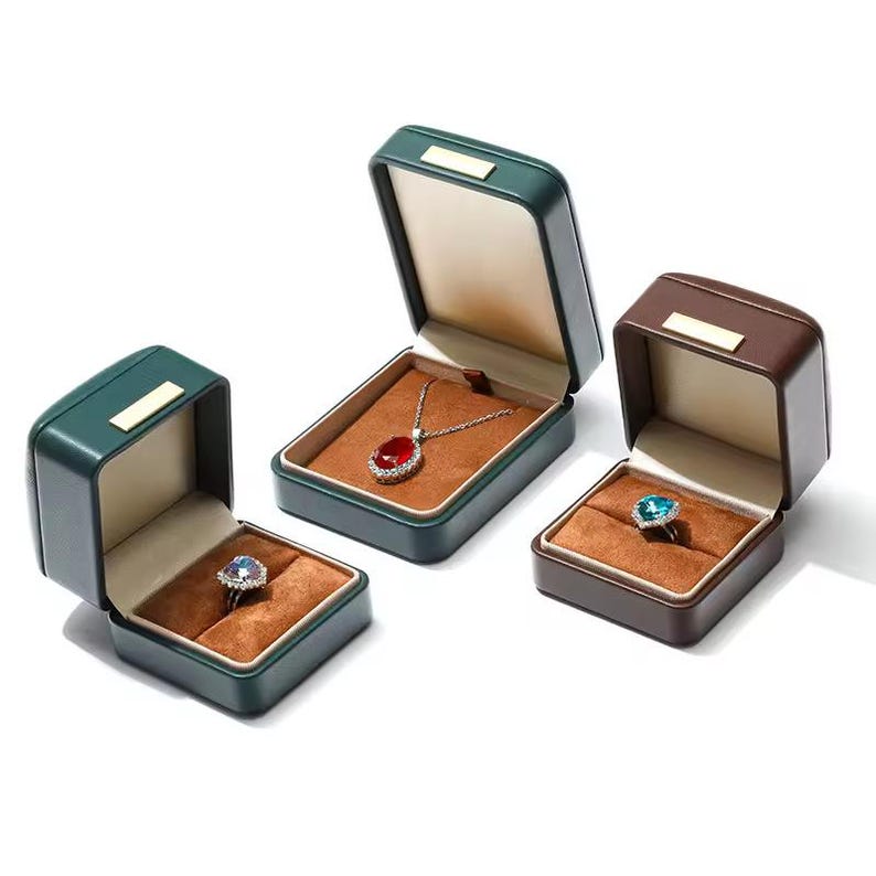 May include: Three green and brown velvet jewelry boxes with lids open. The boxes are lined with brown velvet and contain a red gemstone pendant, a blue gemstone ring, and an empty space.