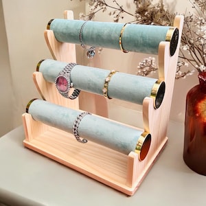 May include: A three-tiered jewelry display stand made of light wood, featuring three cylindrical, light blue velvet-covered bars. Gold-colored metal accents the ends of the bars. Several bracelets and a watch are displayed on the stand.