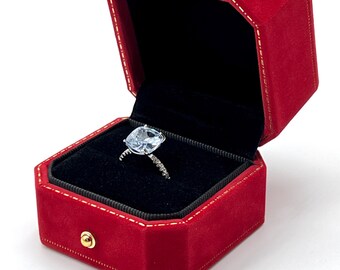 May include: Open red velvet jewelry box with a gold clasp, revealing a silver ring with a large, pear-shaped, light blue gemstone. The ring's band is encrusted with small, clear stones. The box has a black interior.