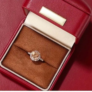 May include: A red leather ring box with a brown velvet interior. A gold ring with a large, round, light brown gemstone is displayed in the box.