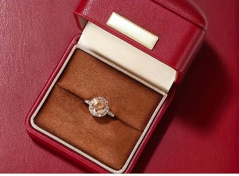 May include: A red leather ring box with a brown velvet interior. A gold ring with a large, round, light brown gemstone is displayed in the box.