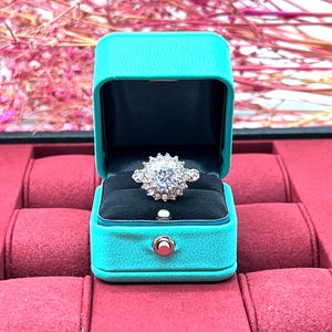 Op de afbeelding: Een sprankelende diamanten ring met een halo-setting ligt in een turquoise ringdoos. De doos staat op een rood fluwelen oppervlak.