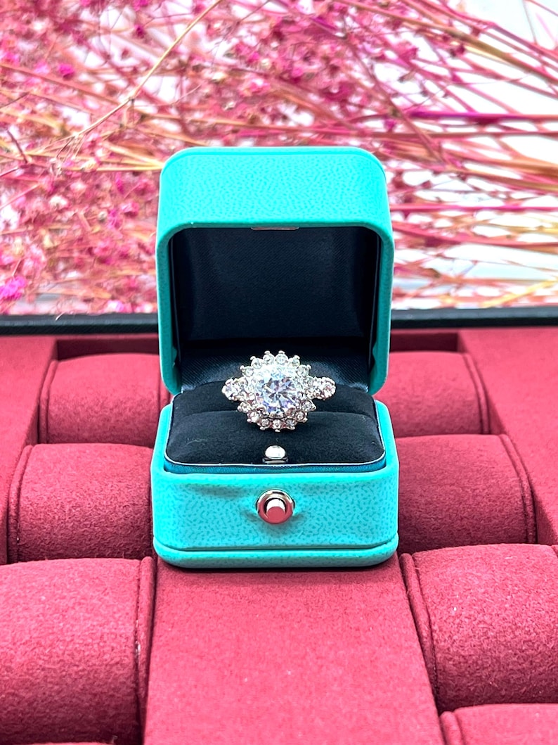Op de afbeelding: Een sprankelende diamanten ring met een halo-setting ligt in een turquoise ringdoos. De doos staat op een rood fluwelen oppervlak.