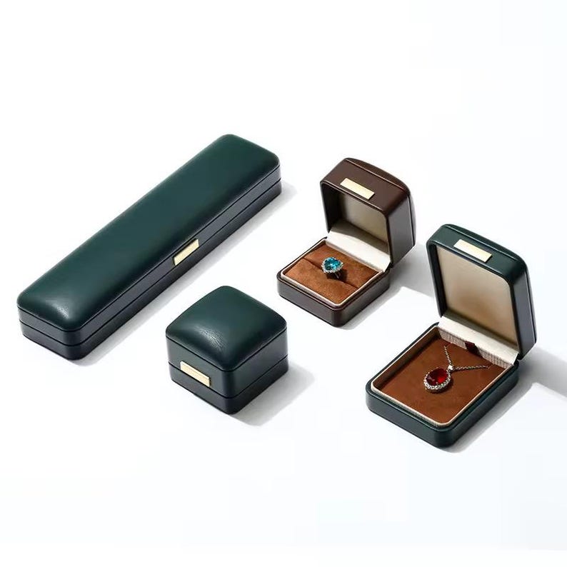 May include: Three green leather jewelry boxes with brown velvet interiors. The boxes are different sizes and shapes. One box is rectangular and has a hinged lid. Another box is square and has a hinged lid. The third box is rectangular and has a sliding lid. The boxes are empty.