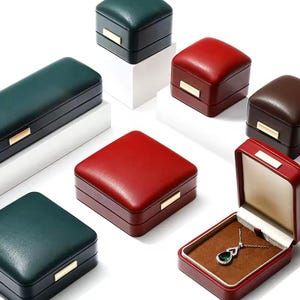 May include: Assortment of small, square, leather-covered boxes in green, red, and brown. The boxes have a hinged lid and are ideal for storing small items like jewelry. One box is open and contains a silver necklace with a green gemstone pendant.