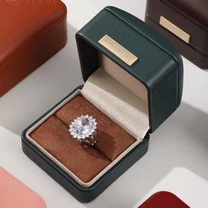 May include: A green velvet ring box with the words "Forever Love" on the lid. The box is open and a ring with a large, clear stone is inside.