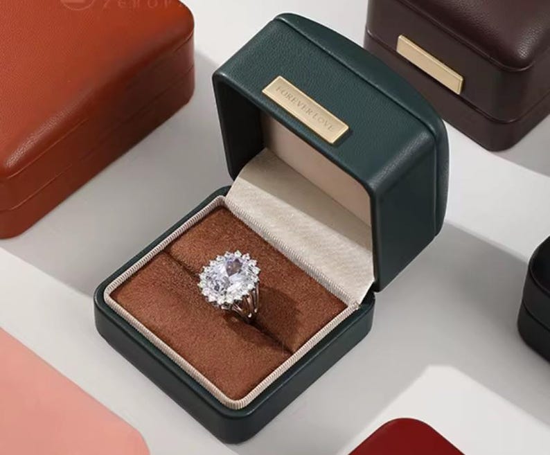 May include: A green velvet ring box with the words "Forever Love" on the lid. The box is open and a ring with a large, clear stone is inside.