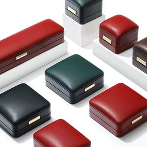 May include: Assortment of six different sized and colored leather jewelry boxes. The boxes are arranged on a white surface. The boxes are all closed and have a simple design.