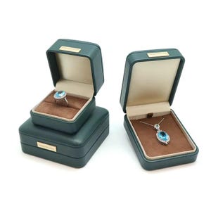 May include: Two green velvet jewelry boxes with brown velvet interiors. One box contains a ring with a large blue gemstone, the other contains a necklace with a pendant featuring a blue gemstone. Both boxes have the text "Forever Love" on the front.