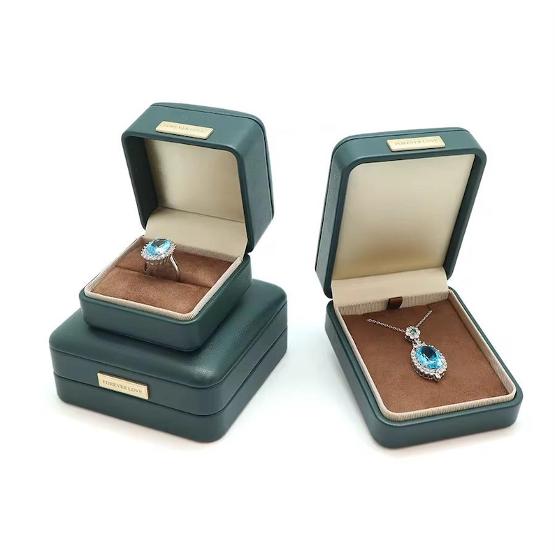 May include: Two green velvet jewelry boxes with brown velvet interiors. One box contains a ring with a large blue gemstone, the other contains a necklace with a pendant featuring a blue gemstone. Both boxes have the text "Forever Love" on the front.