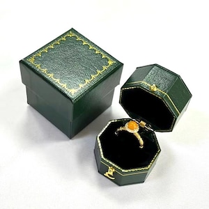 May include: A green ring box with a gold trim and a black velvet interior. The box is open and a gold ring with an orange stone is inside.
