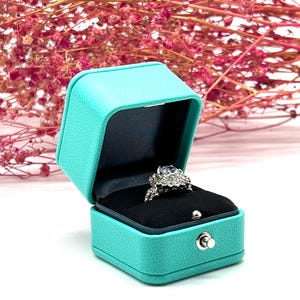 Op de afbeelding: Een turquoise ringendoos met een zilveren ring erin. De ring heeft een grote middelste steen en kleinere stenen eromheen.