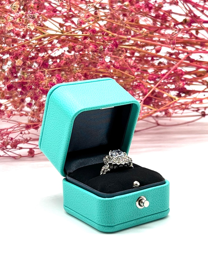 Op de afbeelding: Een turquoise ringendoos met een zilveren ring erin. De ring heeft een grote middelste steen en kleinere stenen eromheen.