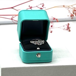 Op de afbeelding: Een turquoise ringdoos met een witte diamanten ring erin. De doos is gemaakt van een zacht, getextureerd materiaal en heeft een scharnierend deksel.