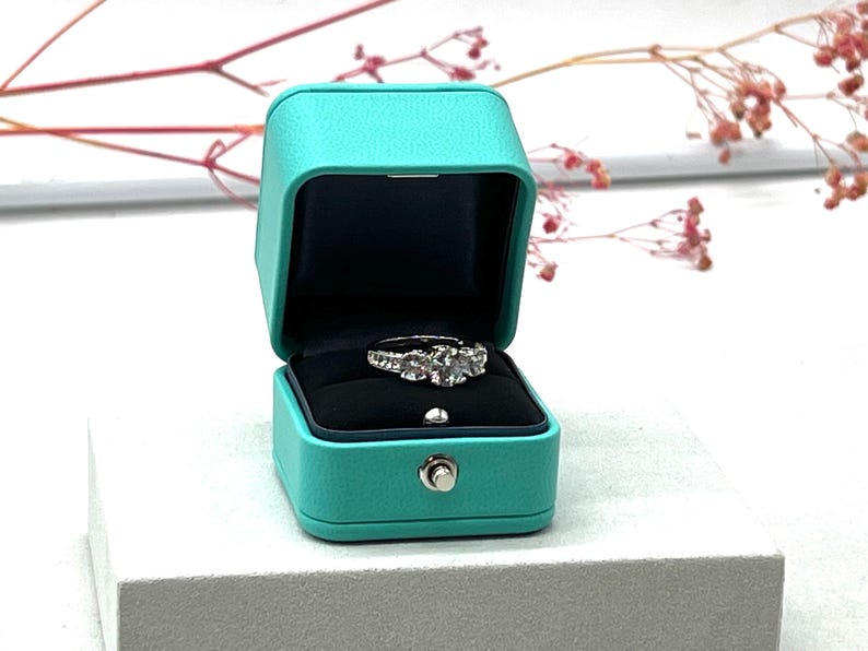 Op de afbeelding: Een turquoise ringdoos met een witte diamanten ring erin. De doos is gemaakt van een zacht, getextureerd materiaal en heeft een scharnierend deksel.