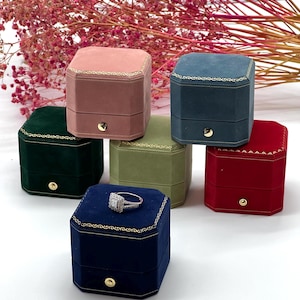 May include: Six velvet ring boxes in different colors: pink, green, blue, gray, red, and navy blue. The boxes are octagonal and have a gold trim. A ring is displayed in the navy blue box.