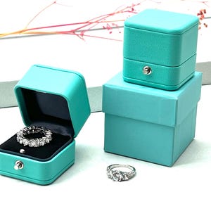 Op de afbeelding: Twee turquoise ringendoosjes, een met een ring erin, en een ring op een wit oppervlak. De ringendoosjes zijn gemaakt van een zacht, fluweelachtig materiaal en hebben een scharnierend deksel. De ring is een zilveren band met een rij kleine diamanten.