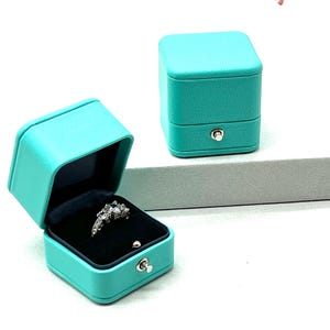 Op de afbeelding: Een turquoise ringendoos met een scharnierend deksel en een ring erin. De doos is gemaakt van een getextureerd materiaal en heeft een zilveren sluiting.