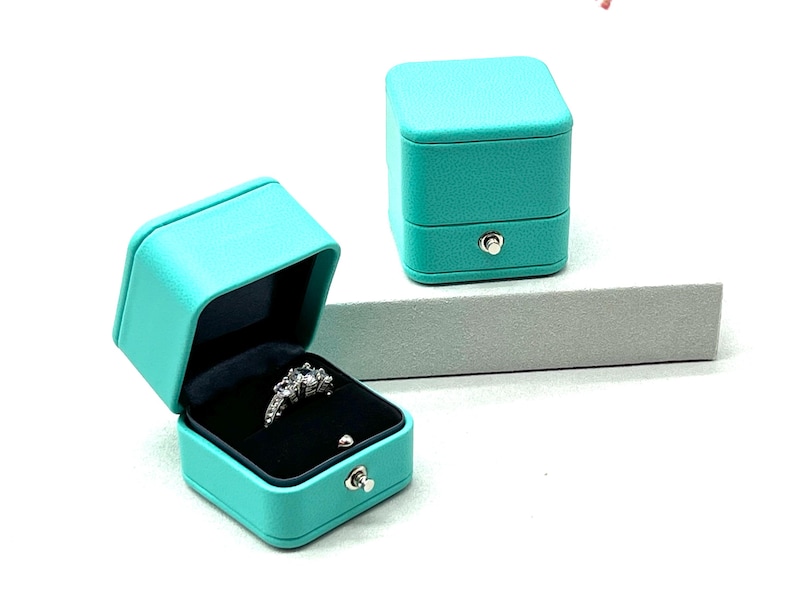 Op de afbeelding: Een turquoise ringendoos met een scharnierend deksel en een ring erin. De doos is gemaakt van een getextureerd materiaal en heeft een zilveren sluiting.