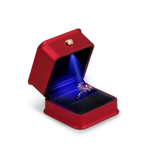 May include: A red jewelry box with a gold clasp, open to reveal a ring with a large, red gemstone. The ring is illuminated by a blue light, set against a black velvet interior.