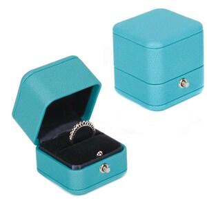 Op de afbeelding: Een turquoise blauwe ringdoos met een scharnierend deksel en een fluwelen interieur. De doos heeft een klein slotje aan de voorkant.