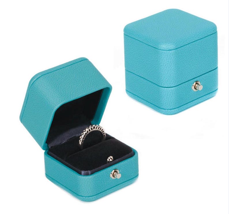 Op de afbeelding: Een turquoise blauwe ringdoos met een scharnierend deksel en een fluwelen interieur. De doos heeft een klein slotje aan de voorkant.