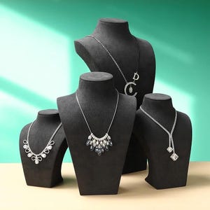 May include: Four black jewelry display busts showcasing various necklaces. The necklaces feature silver chains and sparkling pendants with clear and iridescent stones. The background is a gradient of teal and cream.