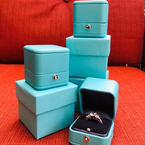 Op de afbeelding: Vier turquoise blauwe ringendozen die op elkaar gestapeld zijn. De bovenste doos is open en bevat een zilveren ring met een heldere steen.