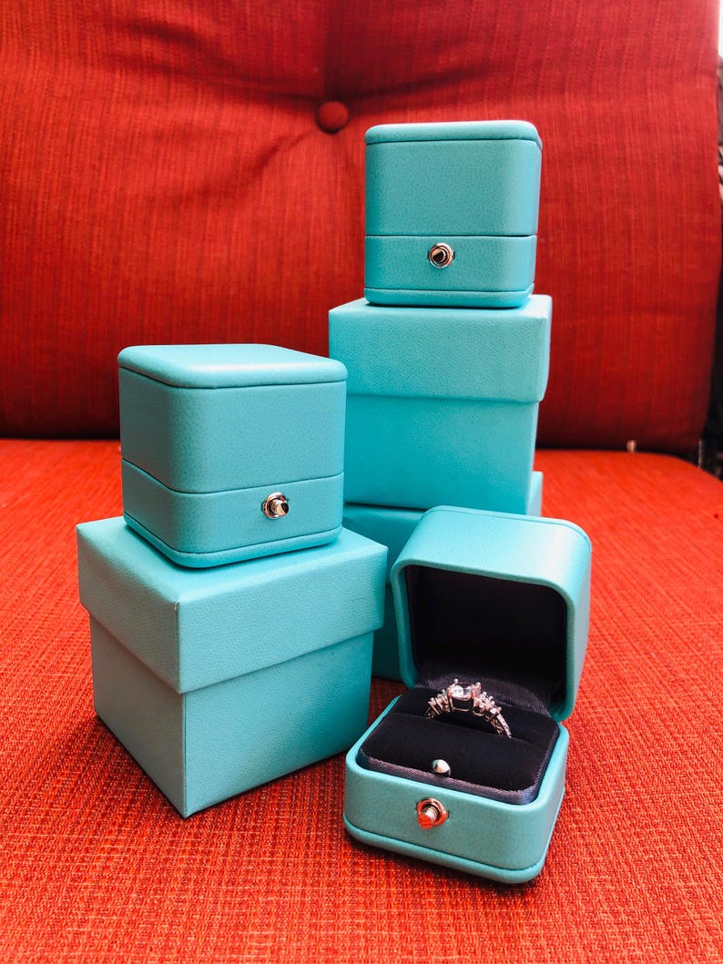 Op de afbeelding: Vier turquoise blauwe ringendozen die op elkaar gestapeld zijn. De bovenste doos is open en bevat een zilveren ring met een heldere steen.