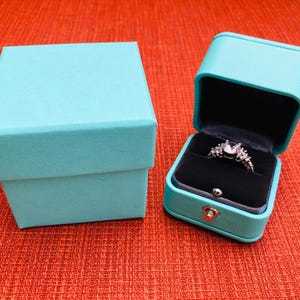 Op de afbeelding: Twee turquoise ringendoosjes. Een doosje is open en bevat een zilveren ring met een grote, heldere steen en kleinere stenen eromheen.
