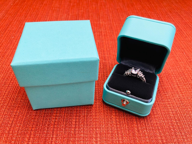 Op de afbeelding: Twee turquoise ringendoosjes. Een doosje is open en bevat een zilveren ring met een grote, heldere steen en kleinere stenen eromheen.