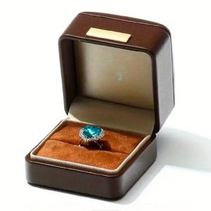 May include: A brown leather ring box with a heart-shaped ring with a blue gemstone and silver setting.