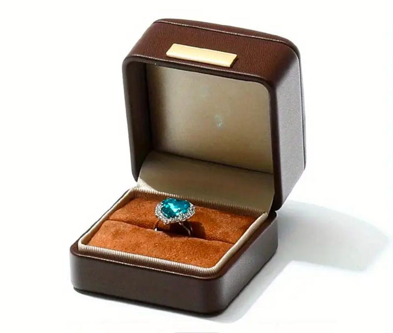 May include: A brown leather ring box with a heart-shaped ring with a blue gemstone and silver setting.