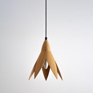 A pendant light fixture with a light wood flower-shaped shade hangs from a black cord against a white background. The shade is made of light brown, possibly oak, wood and has six petals.  The light bulb is visible through the center of the shade. The minimalist design is ideal for modern or contemporary interiors.