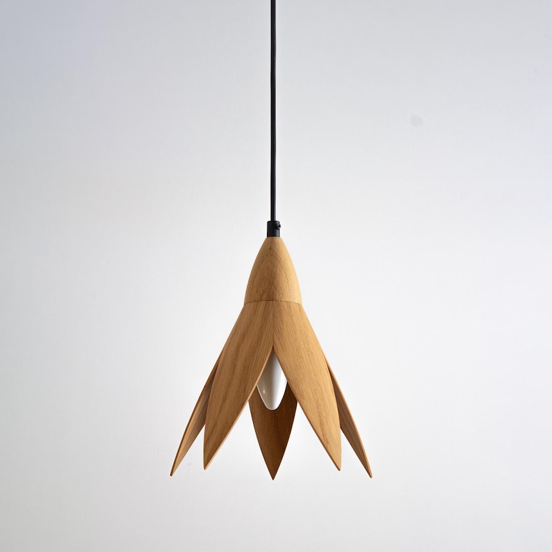 A pendant light fixture with a light wood flower-shaped shade hangs from a black cord against a white background. The shade is made of light brown, possibly oak, wood and has six petals.  The light bulb is visible through the center of the shade. The minimalist design is ideal for modern or contemporary interiors.