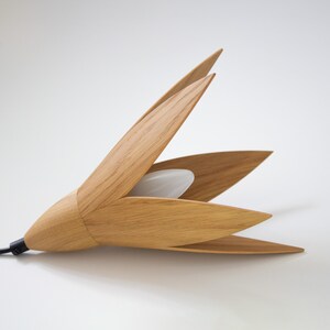 A contemporary wooden pendant light fixture, shaped like a stylized flower with five light oak petals.  The petals are arranged around a central frosted white light source. The sleek design features a minimalist aesthetic, ideal for modern interiors. The light is crafted from natural light oak wood, creating a warm and inviting ambiance. The fixture's simple yet elegant form adds a touch of sophistication to any space. The pendant light is shown lying on a white surface.