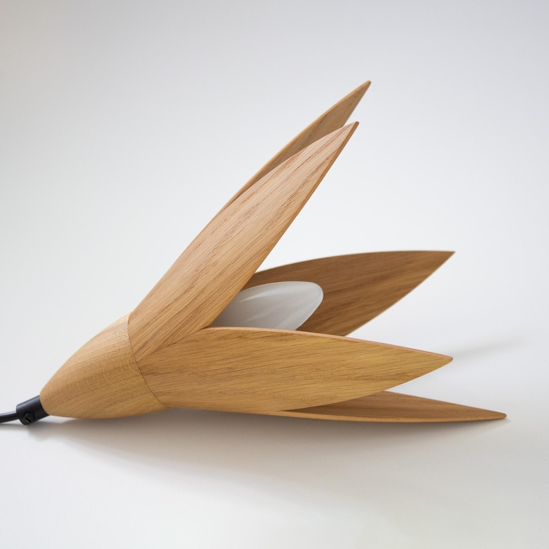 A contemporary wooden pendant light fixture, shaped like a stylized flower with five light oak petals.  The petals are arranged around a central frosted white light source. The sleek design features a minimalist aesthetic, ideal for modern interiors. The light is crafted from natural light oak wood, creating a warm and inviting ambiance. The fixture's simple yet elegant form adds a touch of sophistication to any space. The pendant light is shown lying on a white surface.