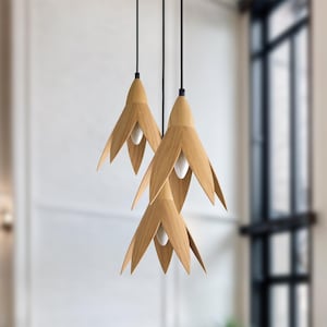 Three modern pendant lights hang from the ceiling. Each light features a light wood, flower-shaped shade with a minimalist design. The lights are suspended by thin black cords, and the overall aesthetic is clean and contemporary.  The warm-toned wood contrasts nicely with the dark cords. These stylish lights would be a beautiful addition to any home decor.