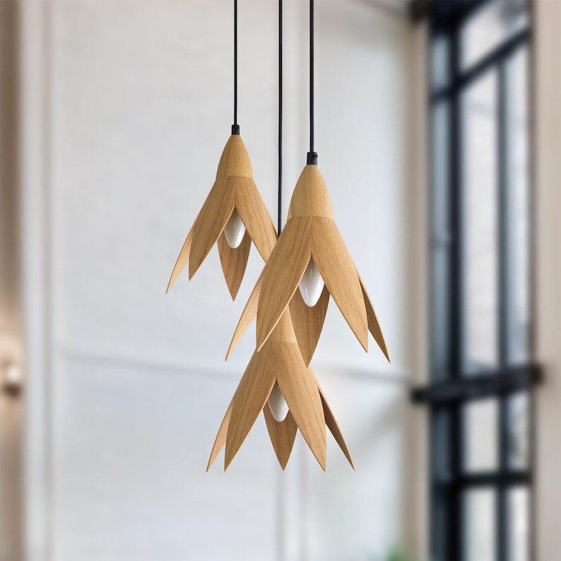 Three modern pendant lights hang from the ceiling. Each light features a light wood, flower-shaped shade with a minimalist design. The lights are suspended by thin black cords, and the overall aesthetic is clean and contemporary.  The warm-toned wood contrasts nicely with the dark cords. These stylish lights would be a beautiful addition to any home decor.