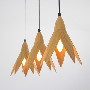 Three modern pendant lights with a minimalist design. Each light features a light brown, wood-like, flower-shaped shade and a black cord. The lights are illuminated, showcasing a warm, ambient glow.  These stylish lights would be a perfect addition to any contemporary home decor.
