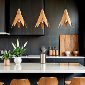 Three modern kitchen pendant lights with a minimalist design. Each light features a light brown, wood-like, tulip-shaped shade hanging from a black cord. The lights are shown hanging above a kitchen counter with dark cabinetry and a white countertop.  The overall style is contemporary and sleek.