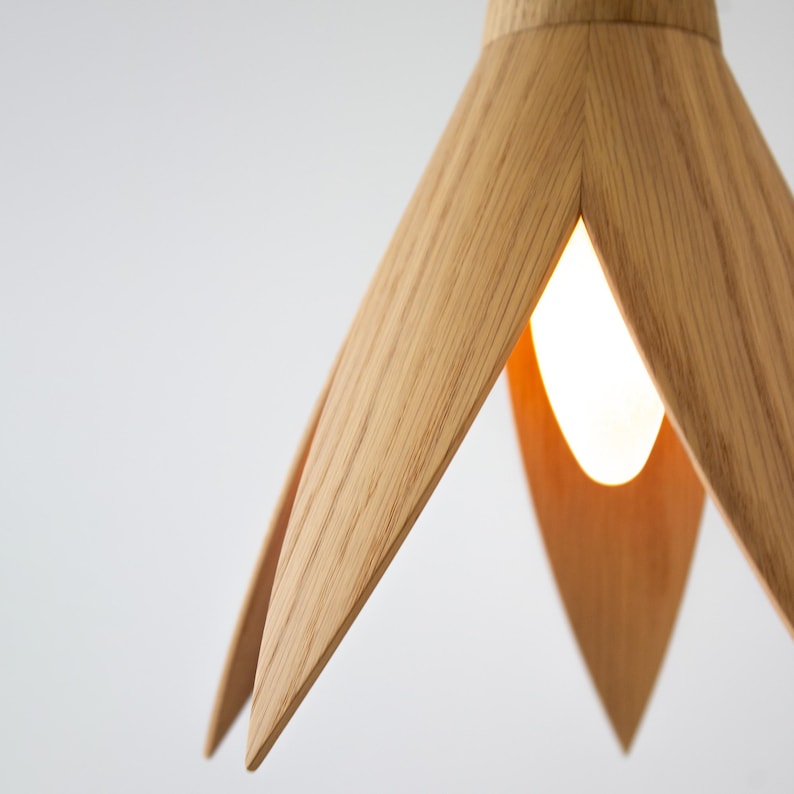 Close-up of a modern pendant light fixture crafted from light brown oak wood. The shade is designed with five slender, petal-like wooden elements that create a unique, flower-like shape.  A warm-toned light bulb is visible through the gaps between the wooden petals, illuminating the interior of the shade. The light's design is minimalist and elegant, suitable for contemporary interiors.