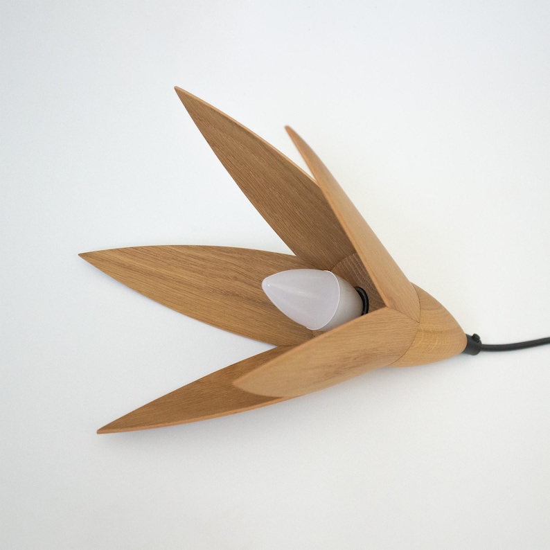 A modern, minimalist pendant light crafted from light brown oak wood. The design features five slender, petal-like wooden pieces radiating from a central point, creating a flower-like shape.  A small, frosted LED bulb is visible within the wooden structure. The light fixture is suspended by a thin, black cord. The overall aesthetic is clean, simple, and organic.