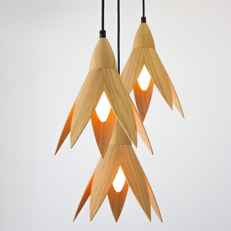 3 wooden pendant lights in the color of natural oak, which in their shape resemble snowdrop flowers, hang on black wires on a white background, warm white bulbs burn in the wooden lamps.